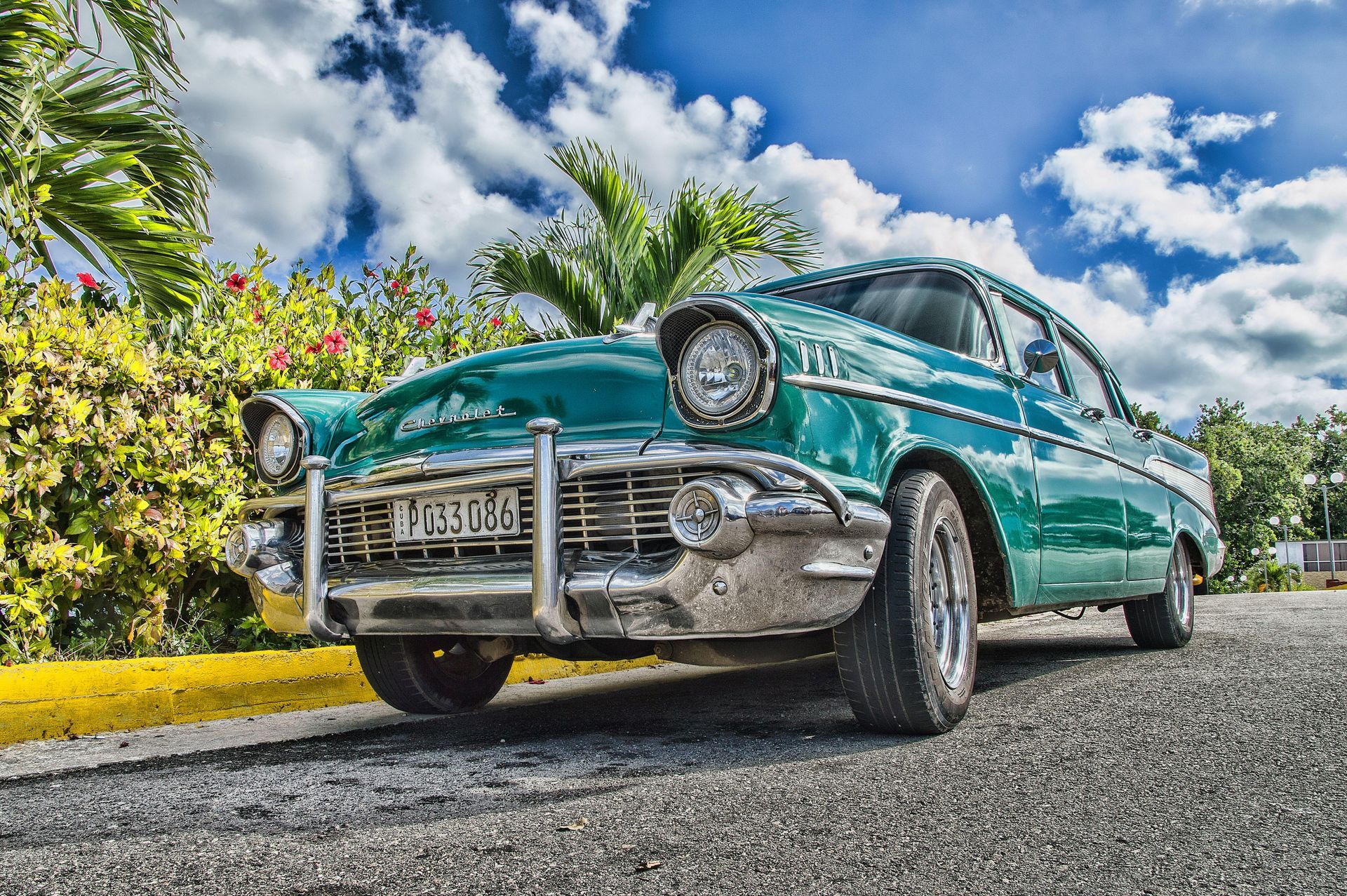 Green classic car parked on a road with a cloudy blue sky and tropical foliage.