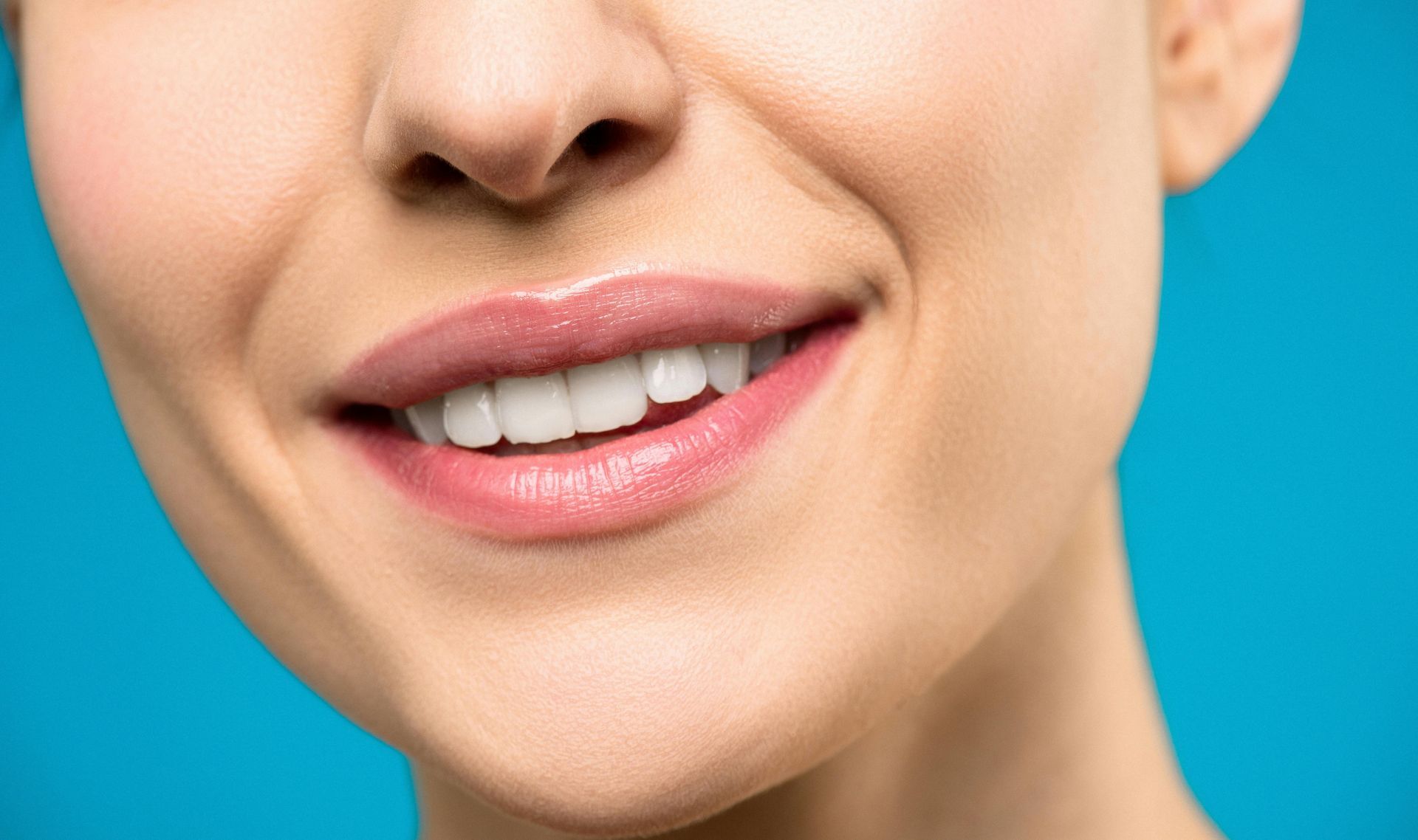Close-up of a person smiling, revealing white teeth and full, pink lips against a blue background.