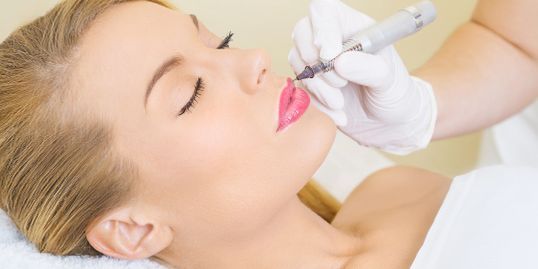 Woman receiving lip tattoo, lying on a white surface with closed eyes, artist using a tattoo machine on her lips.