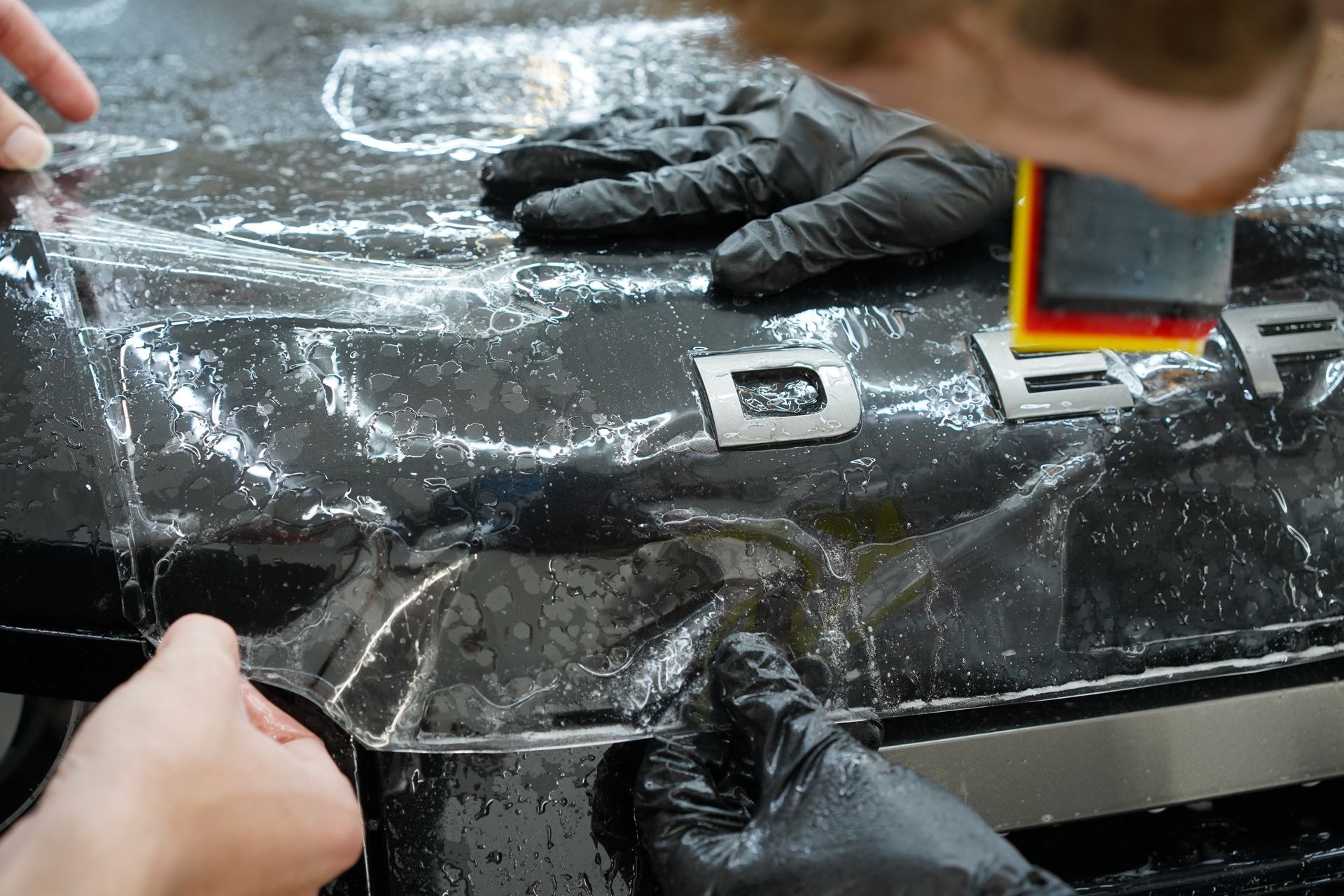 Hands applying clear protective film to the hood of a black vehicle, near silver letters spelling