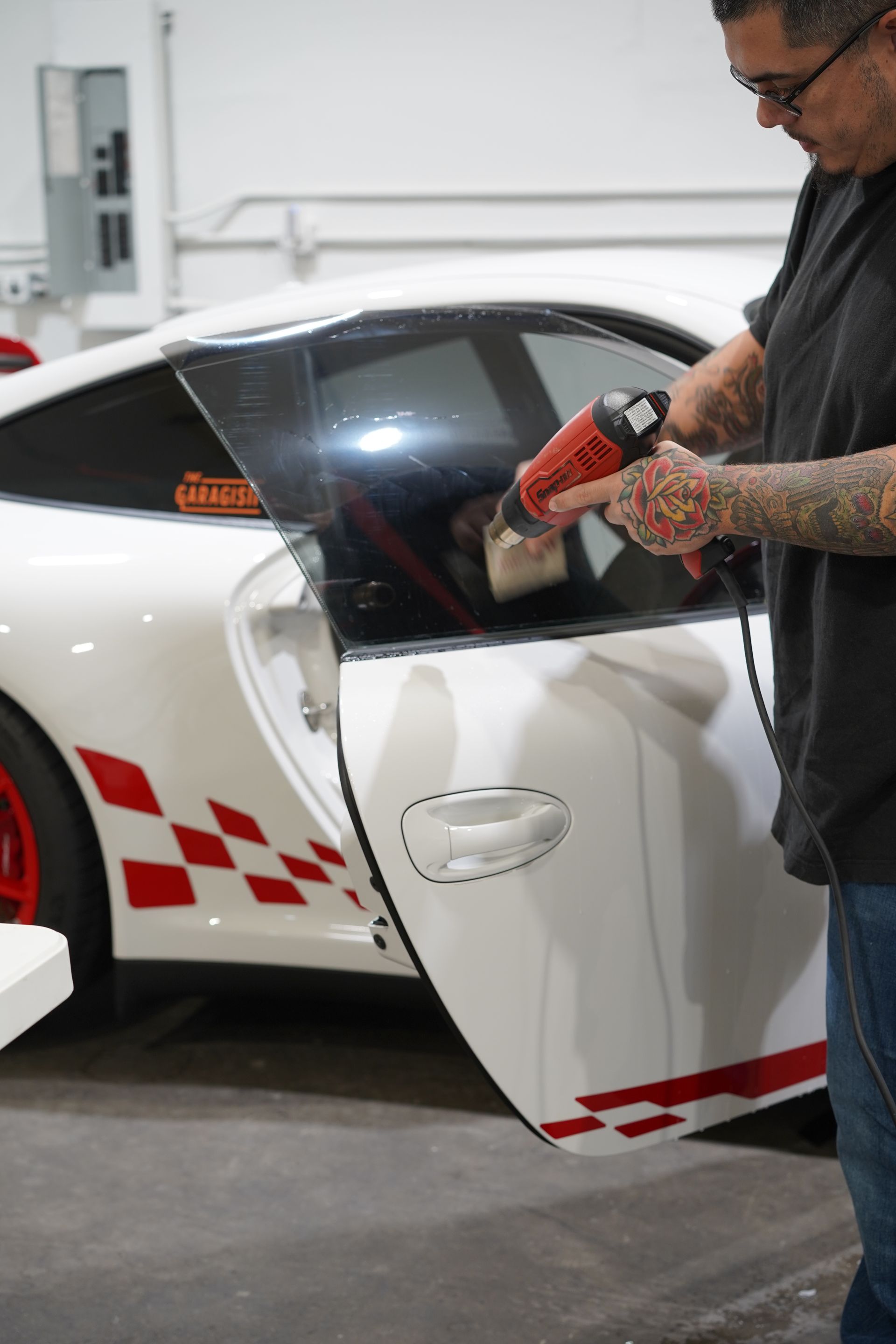 Man applying tint film to a white Porsche window with a heat gun in a garage. The car has red and white checkered decals.