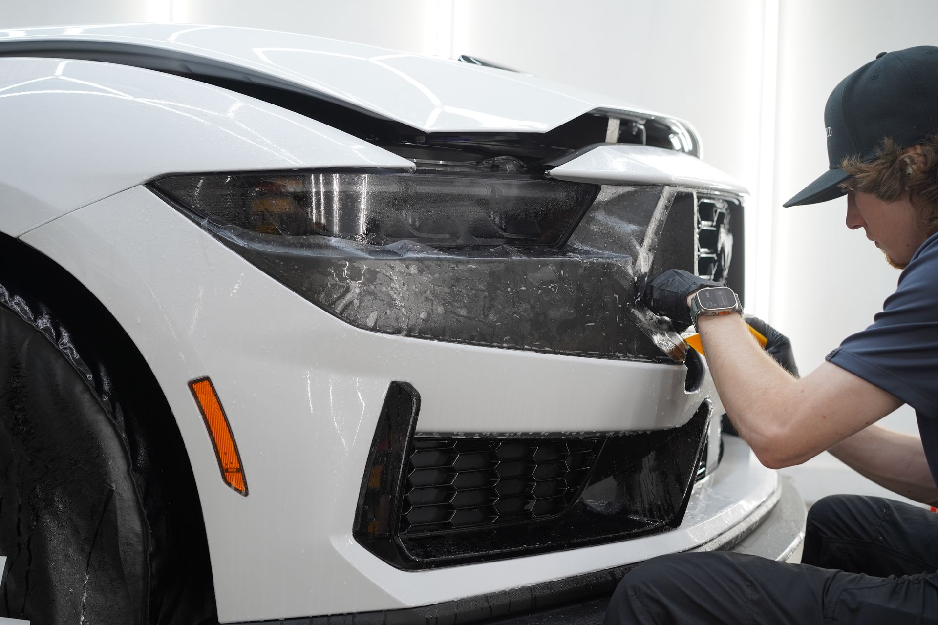 A person applying a clear film to the front of a white car. They are wearing a hat and gloves in a garage-like setting.