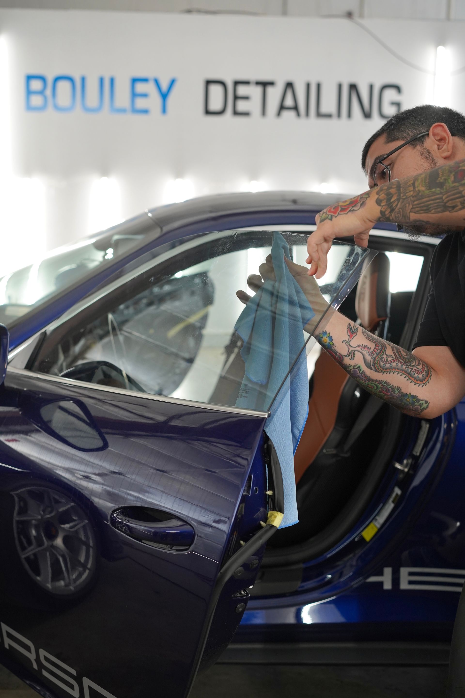 Man applying tint to a blue Porsche window inside a detailing shop. The man has tattoos and is using a towel.