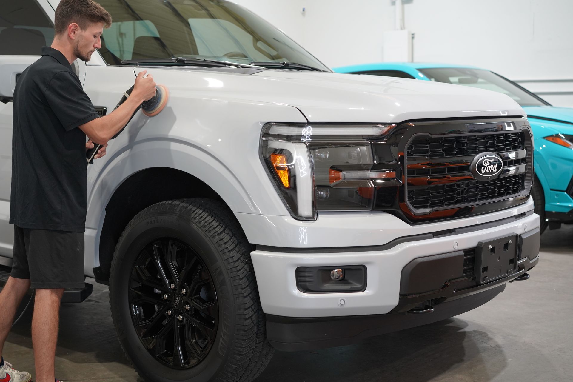A person polishing a light-colored Ford truck. The truck is in a well-lit garage, with a blue car visible in the background.