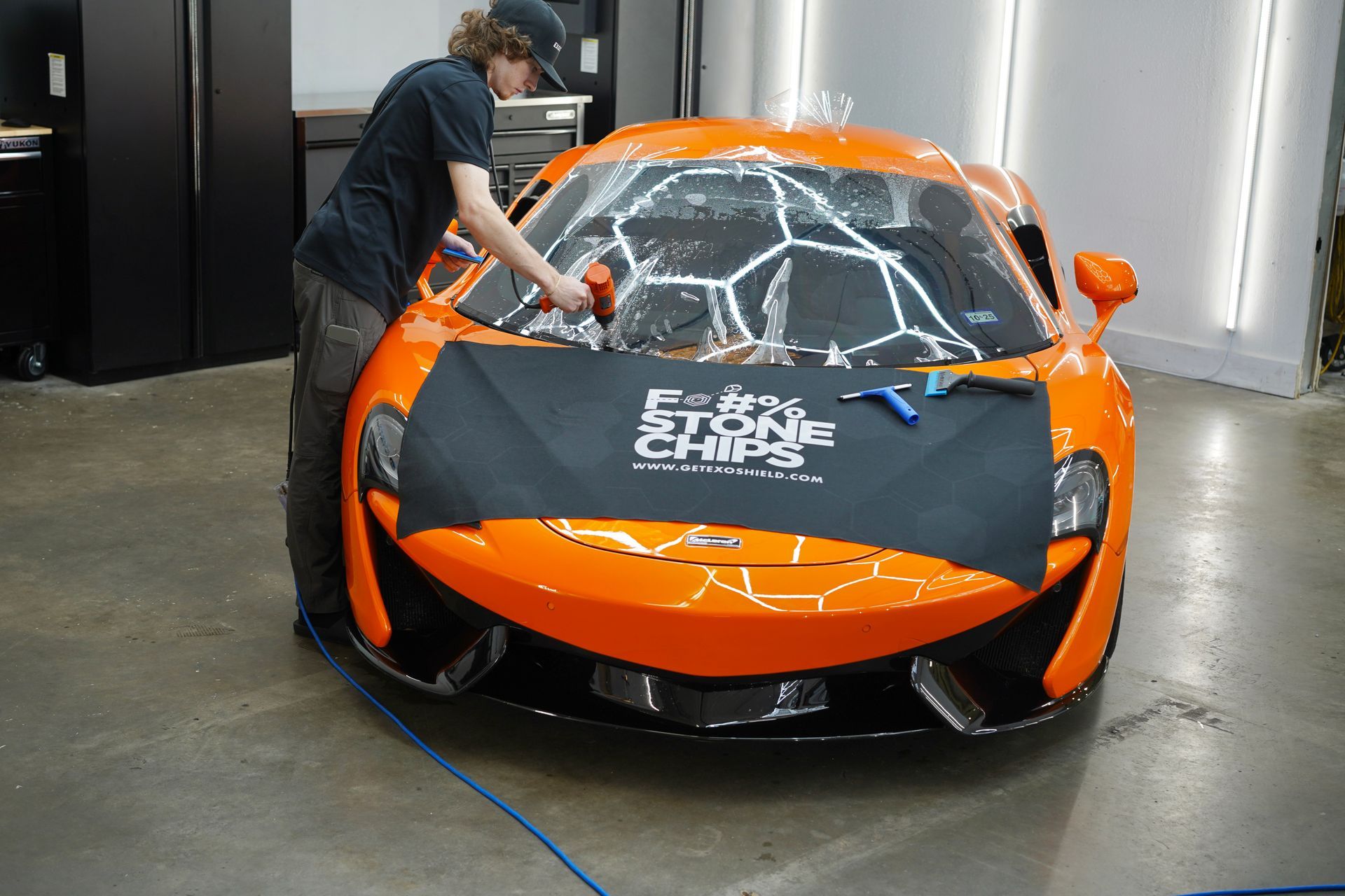 A person applies protective film to the windshield of a bright orange McLaren sports car. The work is being done in a garage.