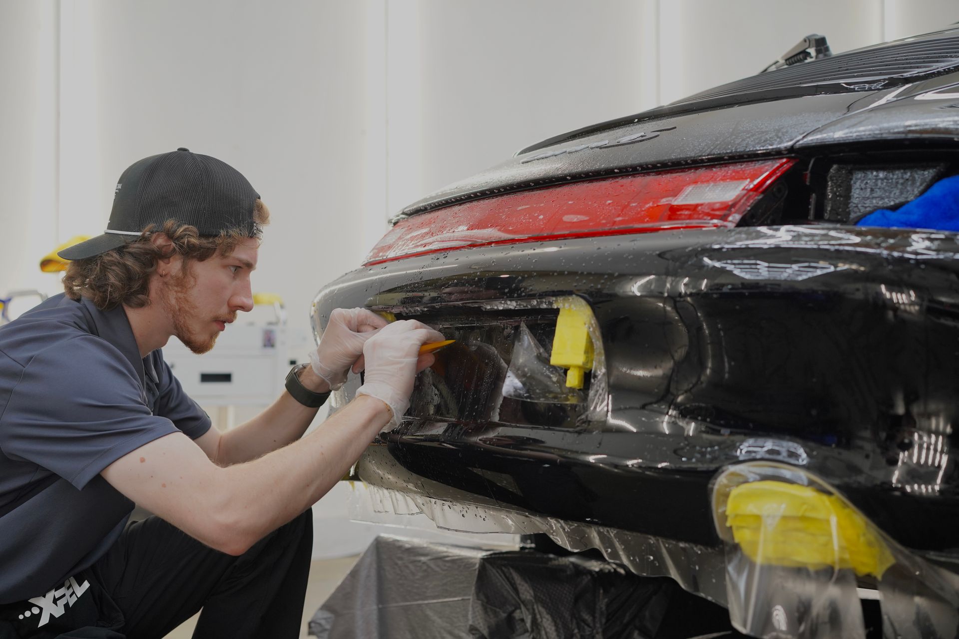 A person installing protective film on the rear bumper of a black car in a garage, using tools.