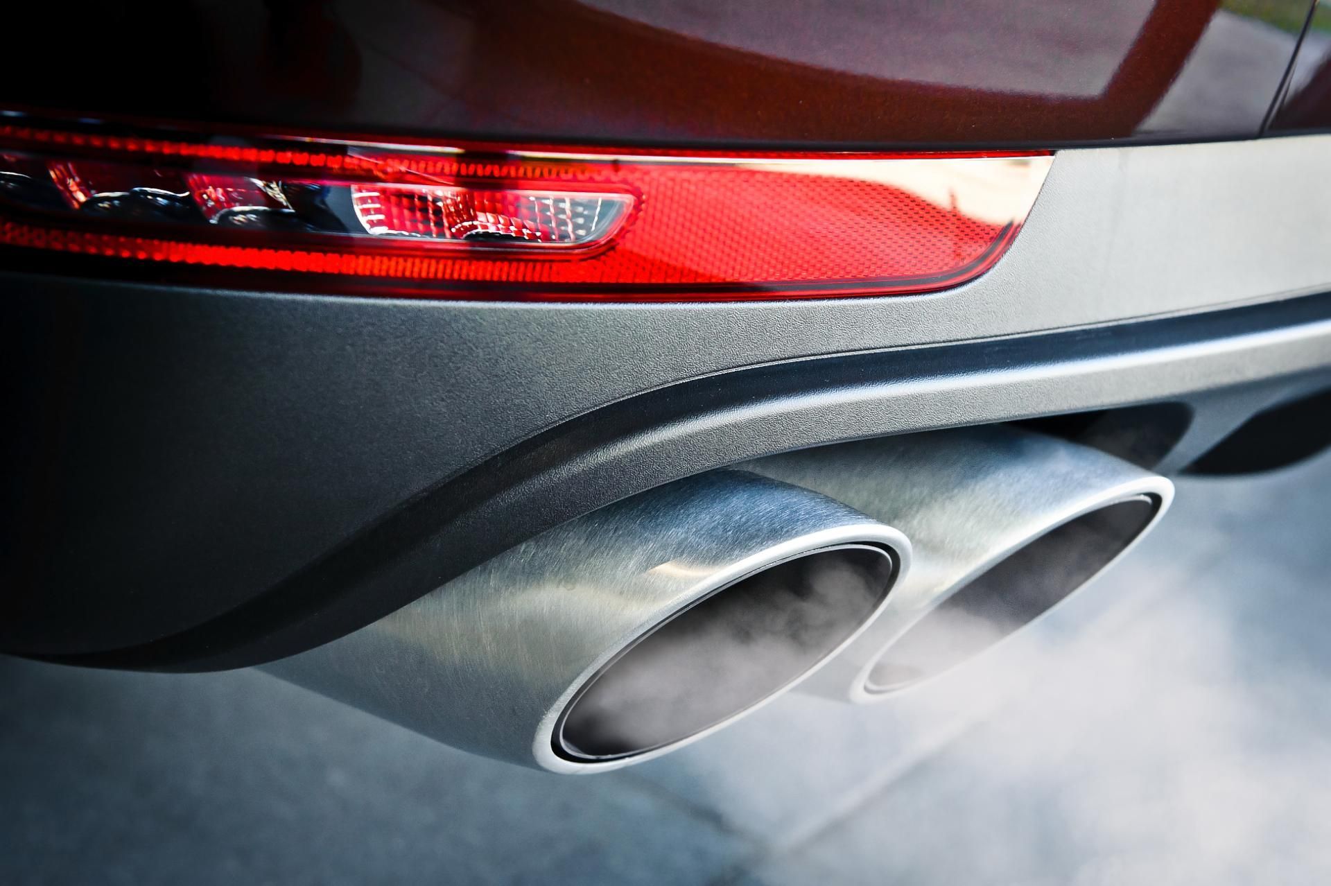 Rear of a gray car with red taillight and dual chrome exhaust pipes releasing smoke