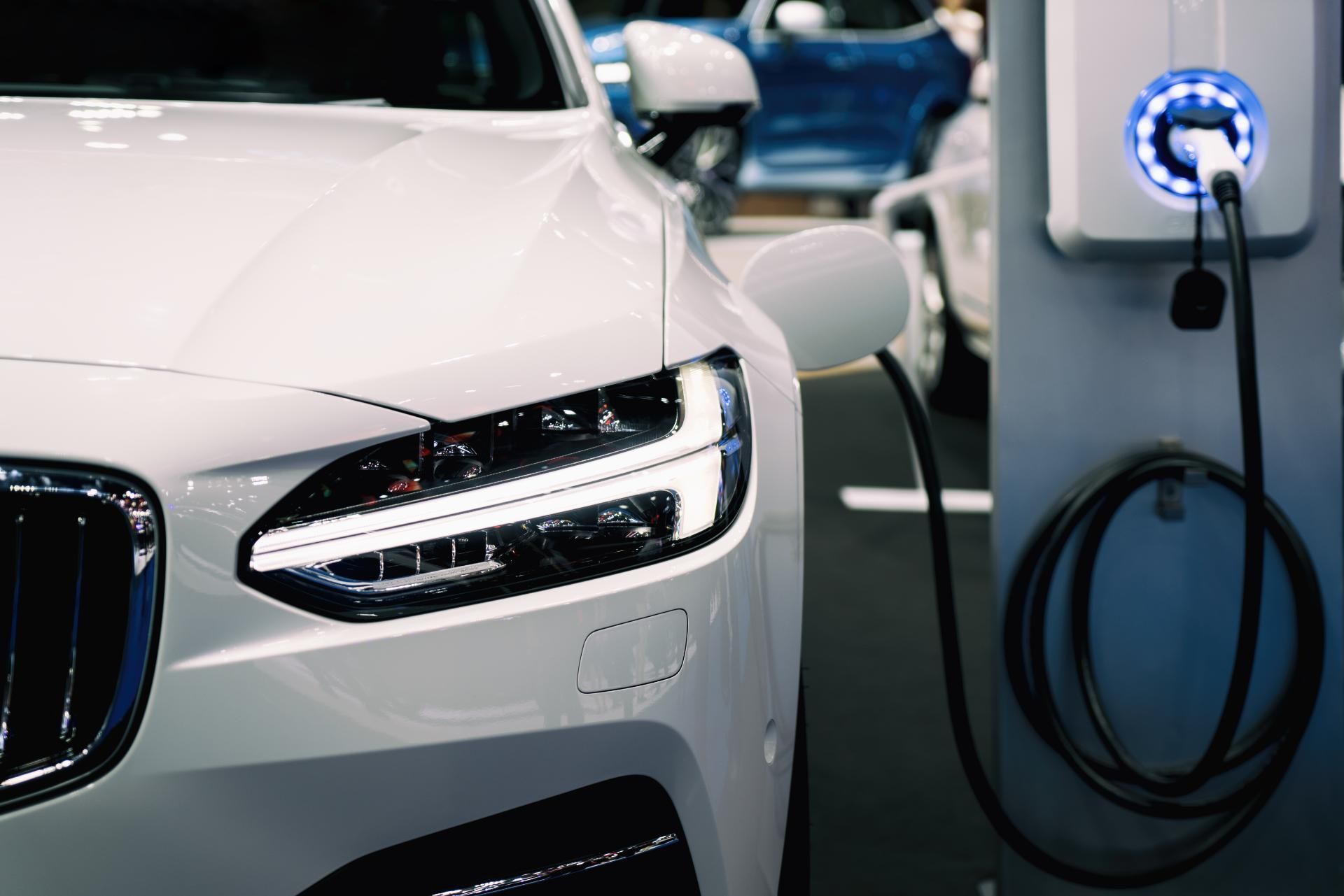White electric car charging at a station, with the front headlight and charging cable visible.