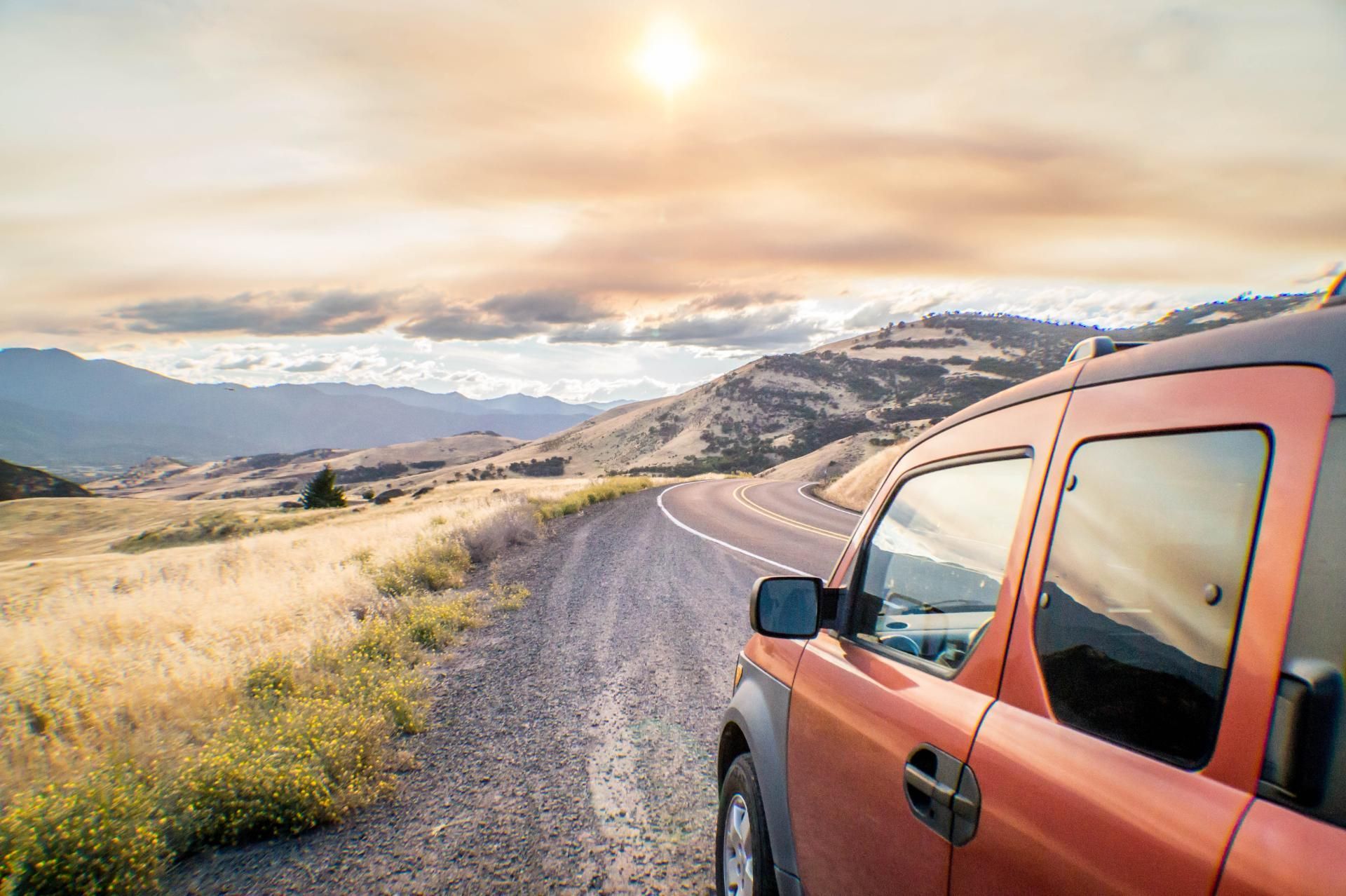 Orange SUV on a gravel road beside mountains at sunset, with wildflowers and a bright sky