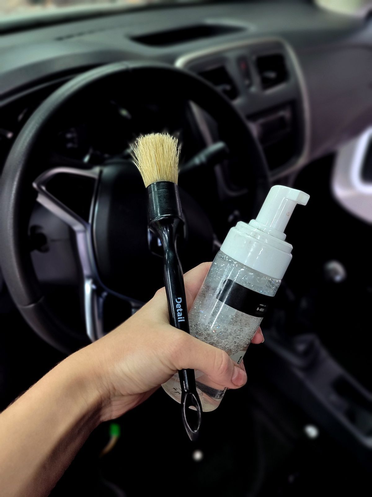 Mano sosteniendo un cepillo y una botella de espuma dentro de un automóvil, listo para detallar.