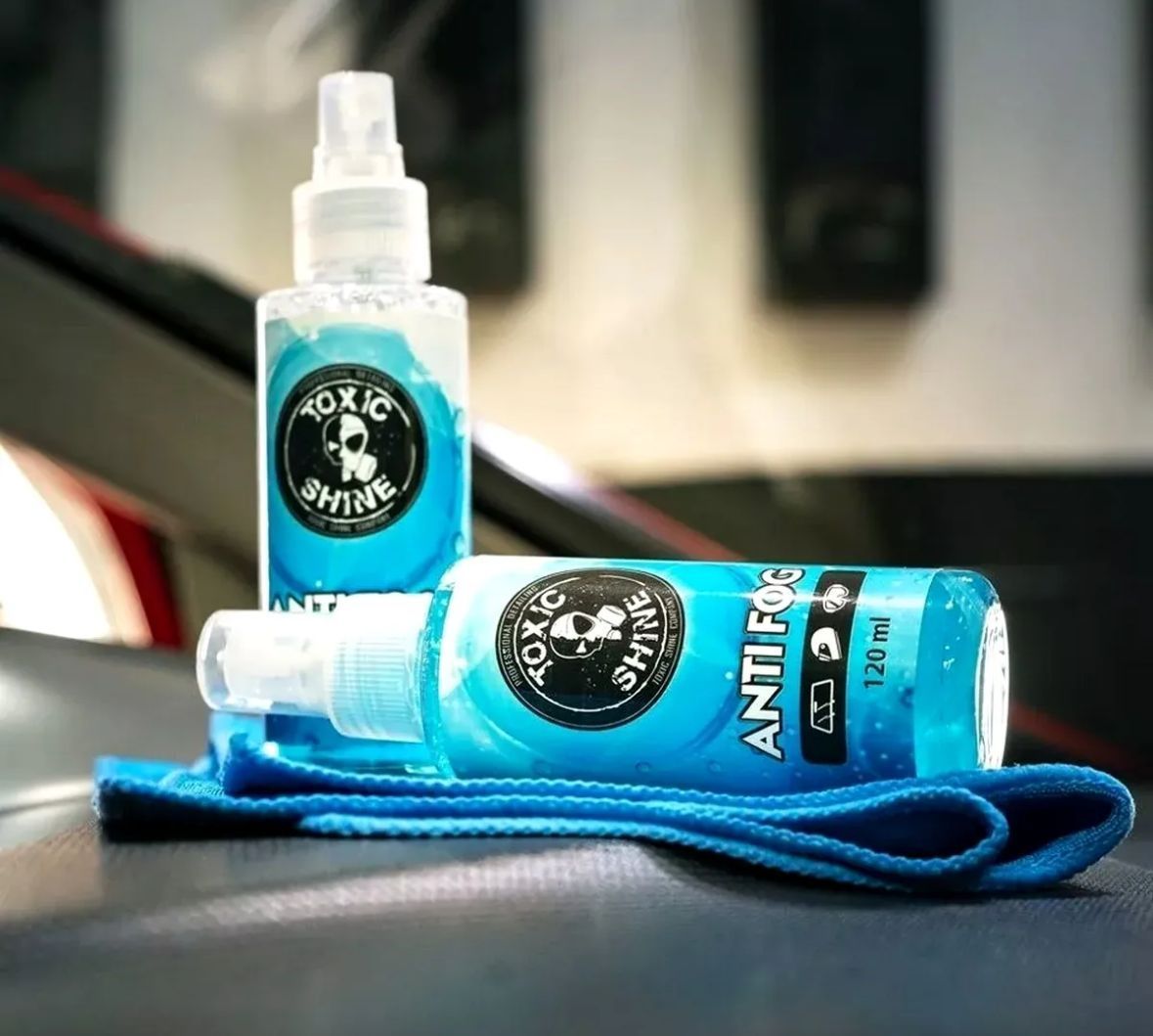 Dos botellas azules de spray "ANTI-NIEBLA" y un paño azul se encuentran sobre un coche, reflejándose en la superficie oscura.