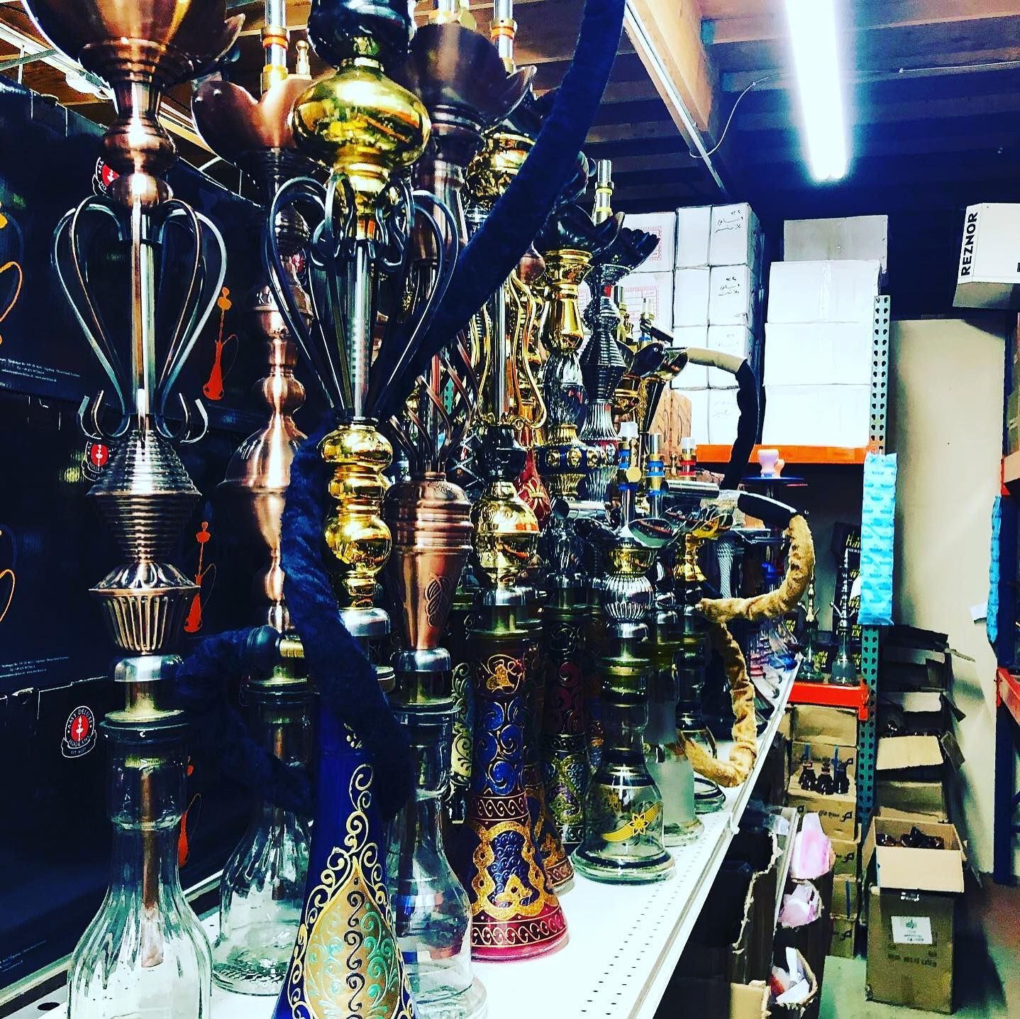 Hookahs displayed on a shelf in a shop. Various colors, tall stems, glass bases. Shelving with storage in the background.