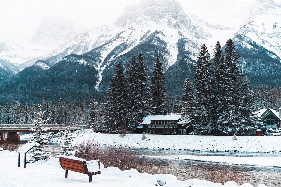 A bench is sitting in the snow near a river with mountains in the background.