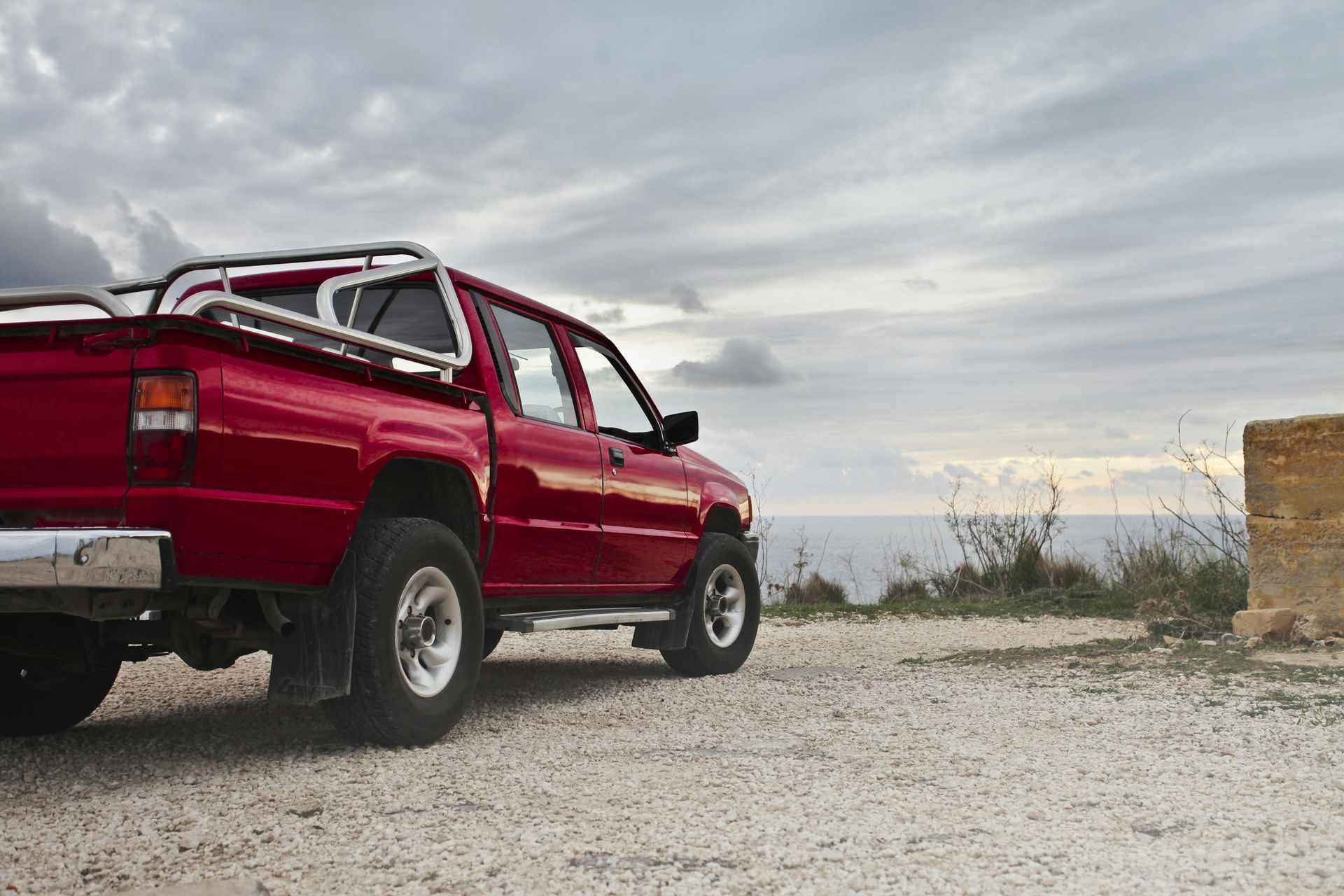 A red truck is parked on a gravel road near the ocean.