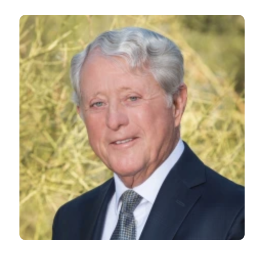 Man in a suit smiles at the camera, with a background of out-of-focus yellow-green vegetation.