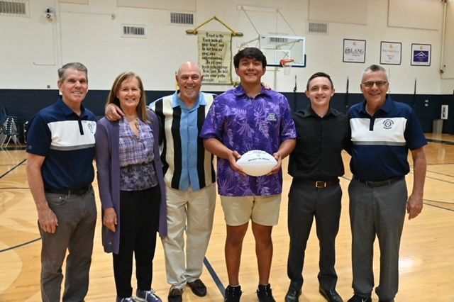 Group poses with a football in a gymnasium. People wear a variety of clothing; one holds the ball.