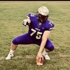 Football player in purple uniform, helmet, and number 75, snapping ball on green field.