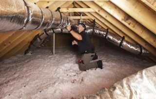 A man is kneeling down in an attic looking at a pipe.