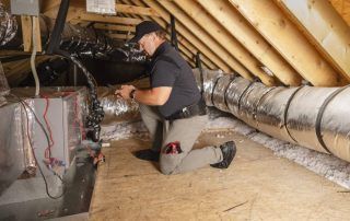 A man is kneeling down in an attic working on an air conditioner.