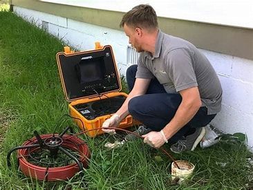 Technician kneels by an open yellow inspection case beside a grassy wall, handling cables and equipment.