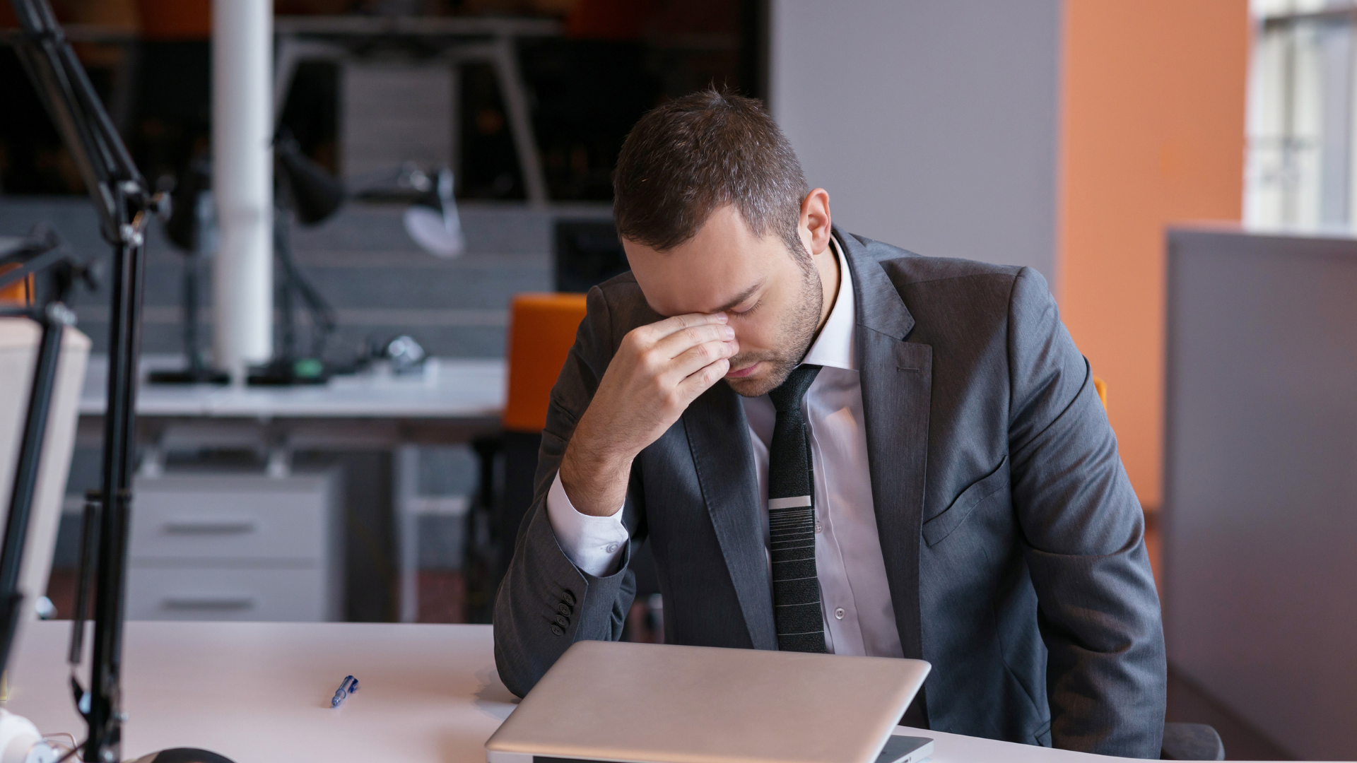 Man in a suit sitting at a desk, holding his forehead and looking stressed in an office.