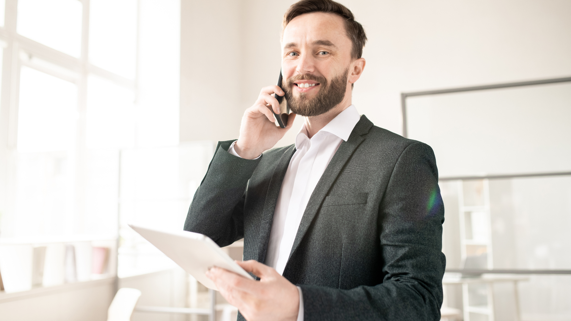 A man in a suit is talking on a cell phone while holding a tablet.