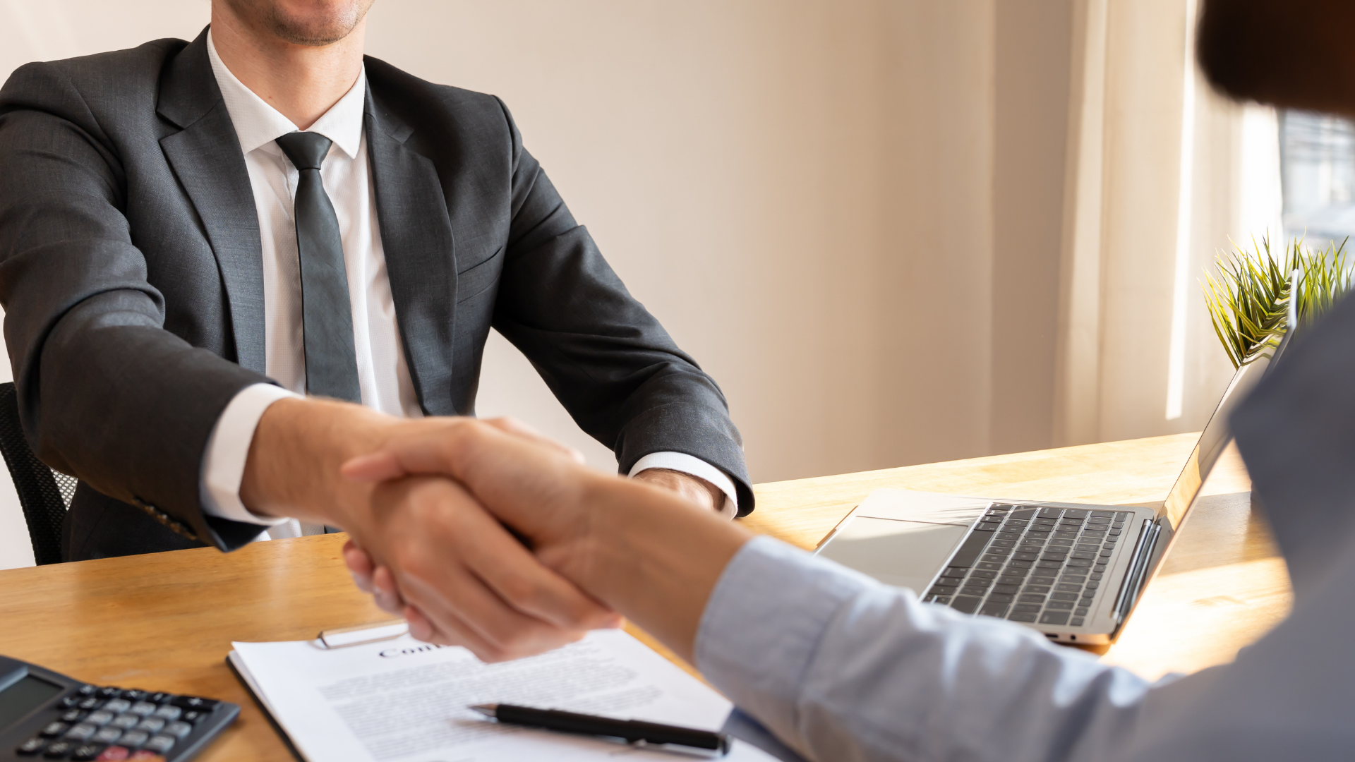 Businesspeople shaking hands across a desk in a bright office with a laptop and documents nearby