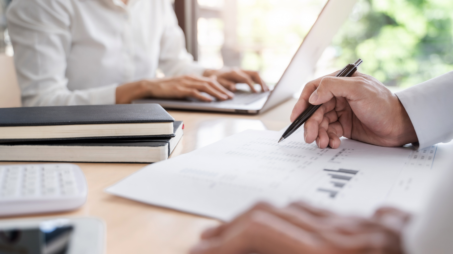 Hands writing notes beside a laptop and stacked books in a bright office meeting setting