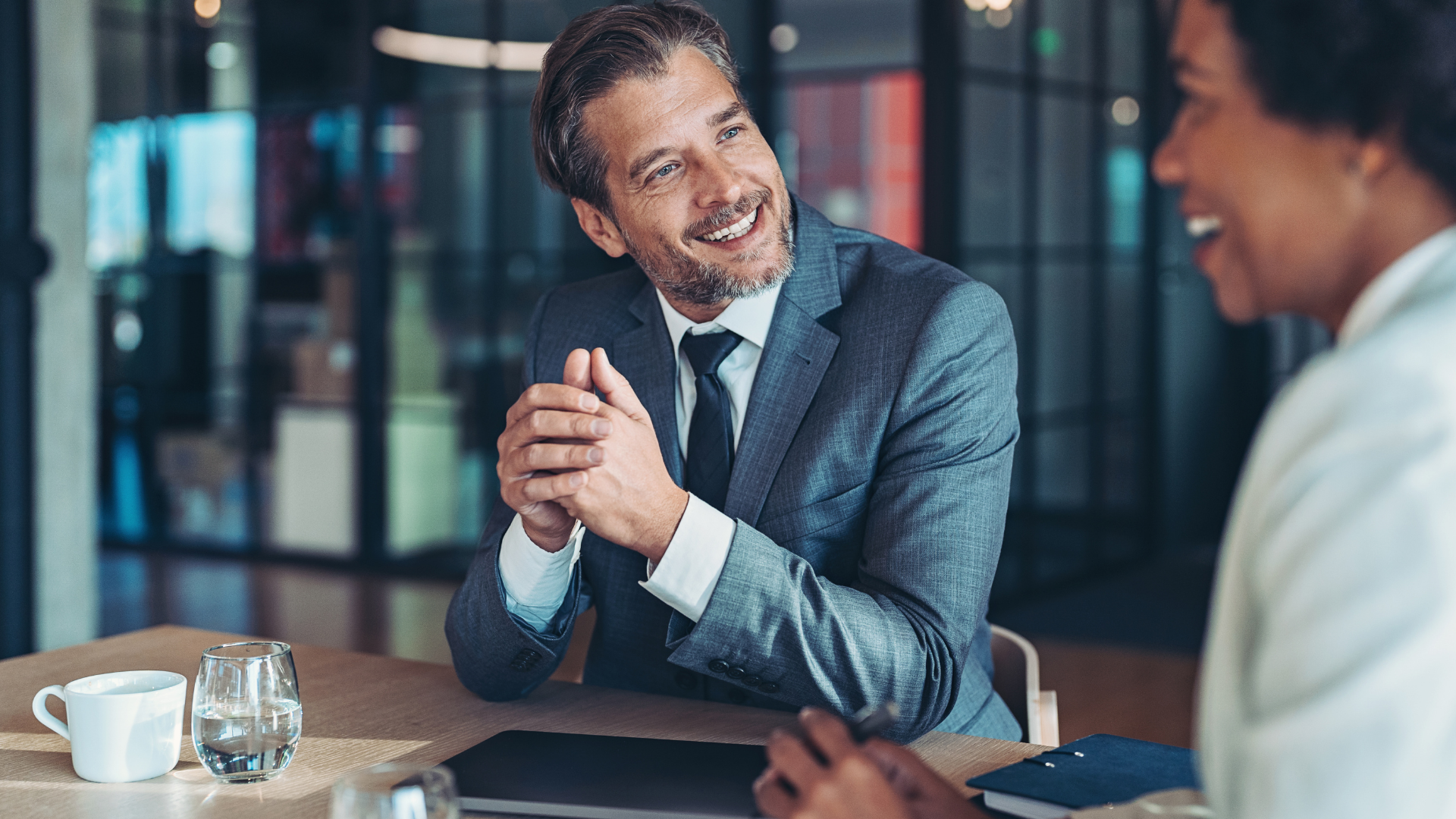 Two coworkers smiling and talking at a table in a modern office setting