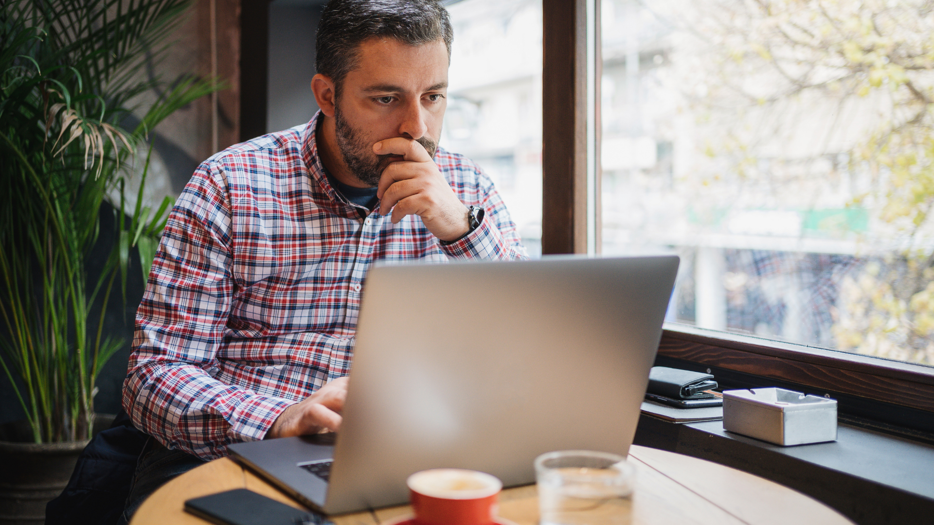Man sitting at a table with a laptop, looking thoughtful in a bright café or office setting.