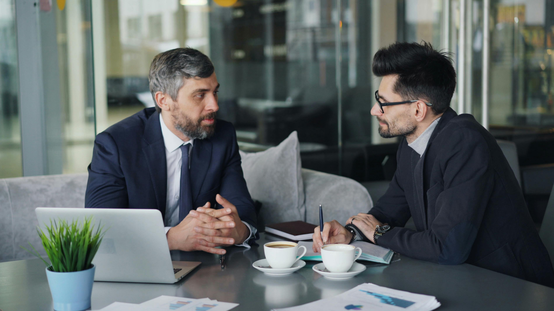 Two professionals in suits talking at a table with a laptop and coffee cups in a modern office setting