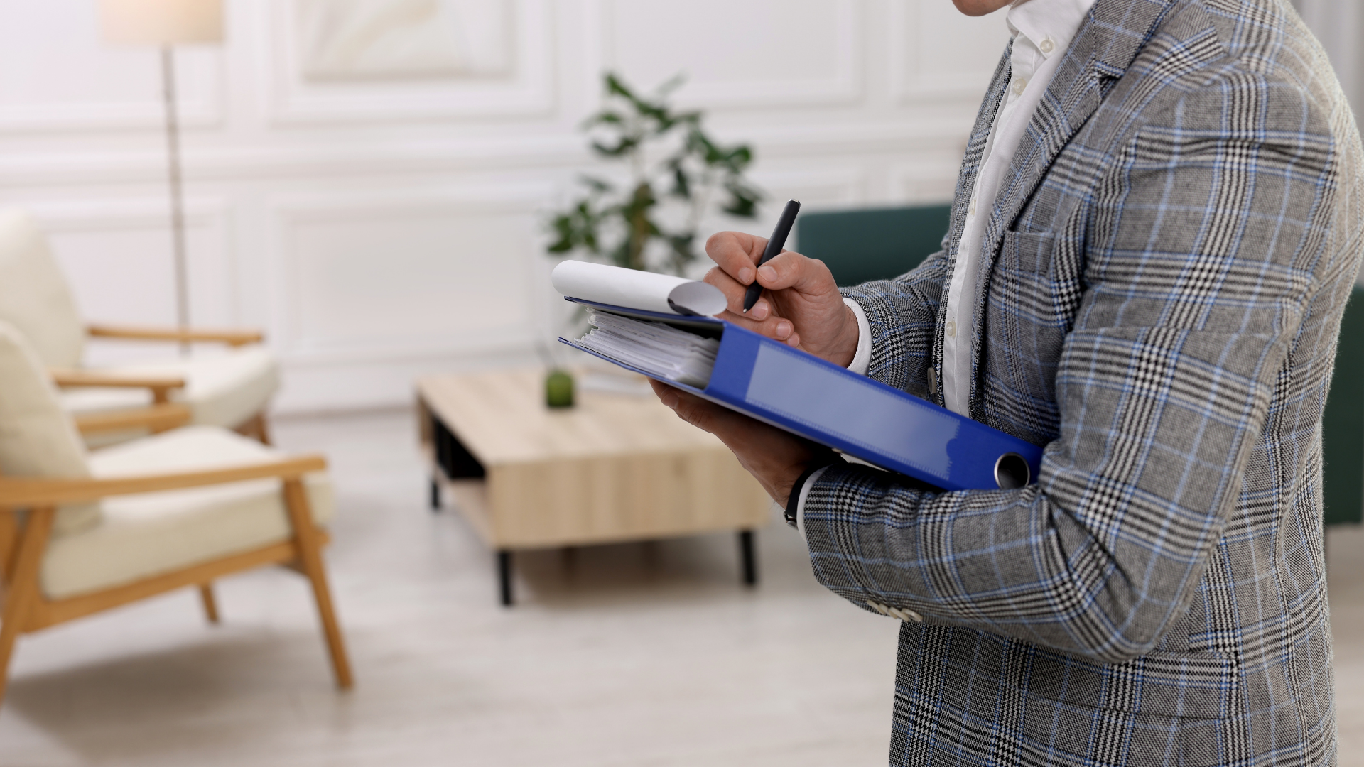 Person in gray blazer writing on documents in a blue binder in a living room.