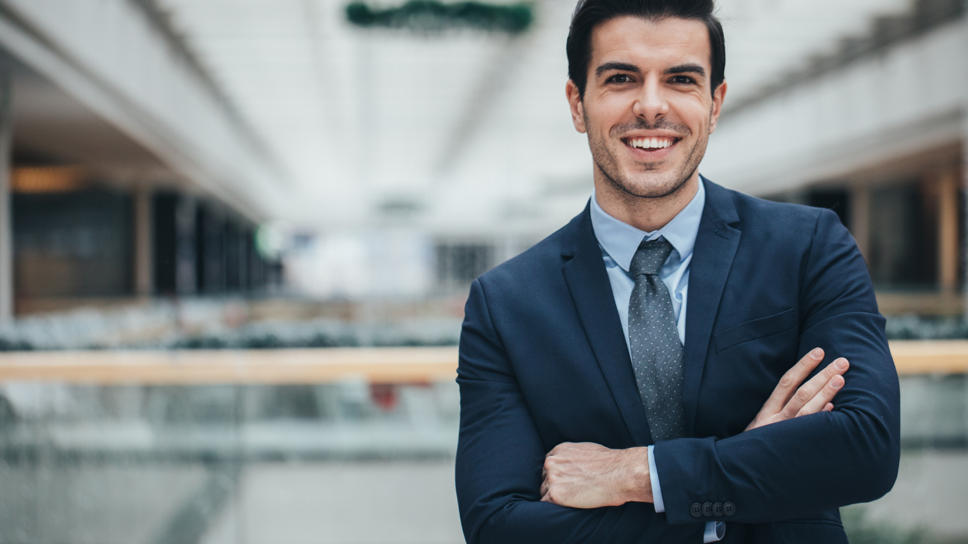 Man in a navy suit, arms crossed, smiling broadly in a modern office.