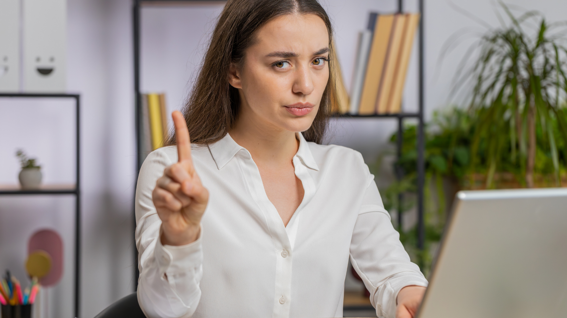 Woman in white shirt gestures with index finger, looking sternly at laptop, possibly in office.