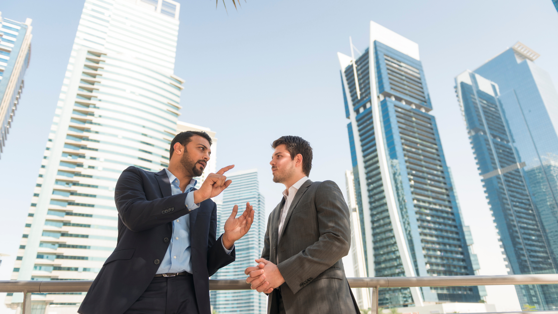 Two men in suits talking outside, tall buildings in background. One gestures and points.
