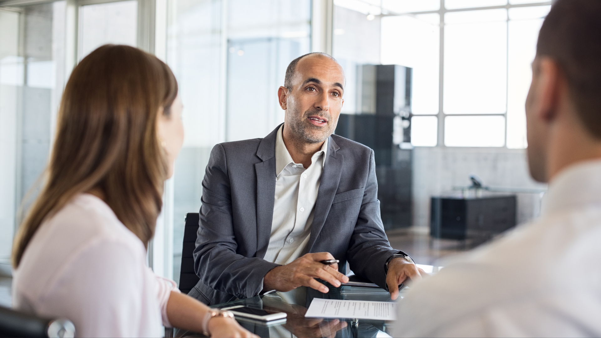 A man in a suit speaks to a couple seated at a table in an office. They appear to be discussing paperwork.