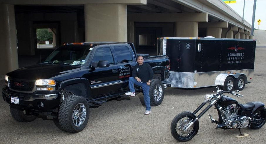 Man sits on a black pickup truck, with a trailer and chopper motorcycle parked nearby under an overpass.