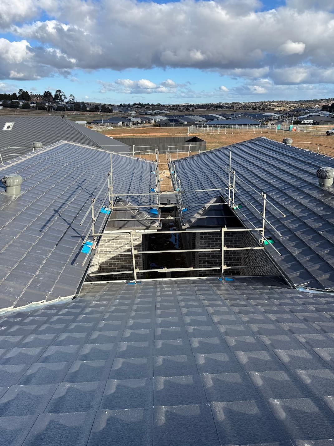 Scaffolding frames an open section between two grey tiled roof slopes on a sunny day with clouds.