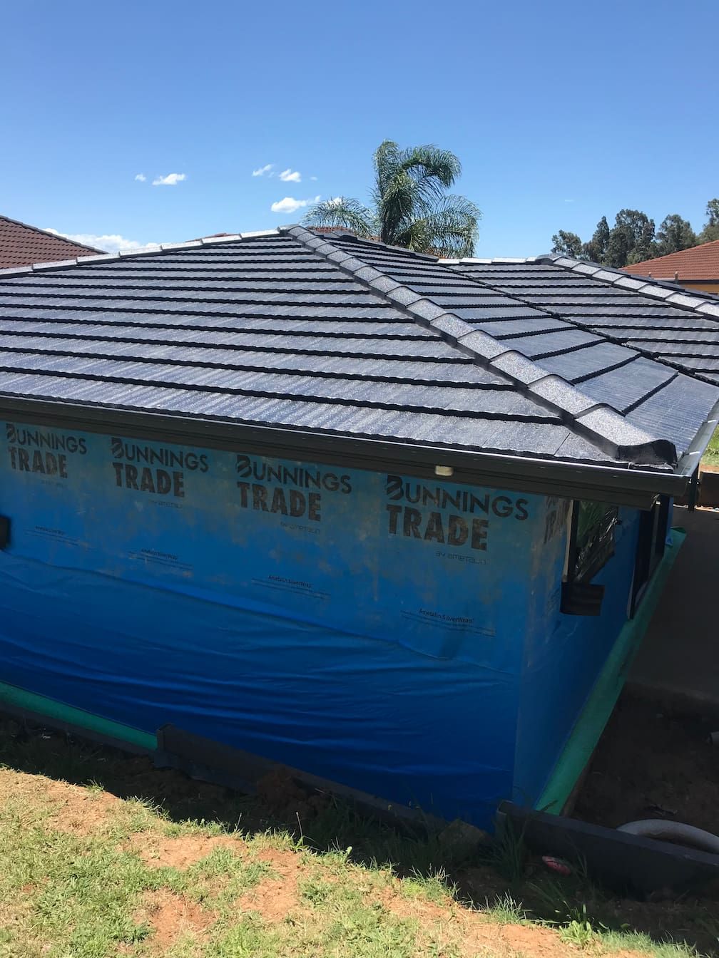 A small building structure under construction with a gray tiled roof and blue protective siding wrap, set on a grassy hill.