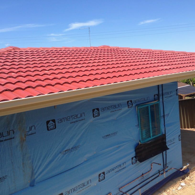 A house under construction with a red tiled roof and blue exterior wrap, showing a small window and exposed pipes.