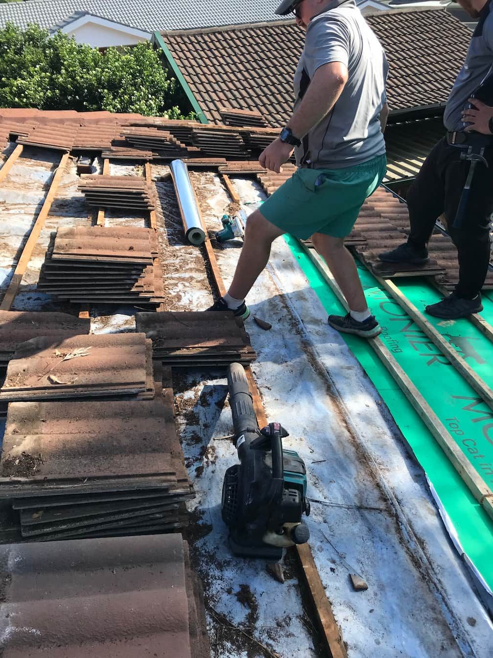 Two workers in shorts and athletic shoes stand on a roof undergoing repairs, with stacks of old tiles and tools nearby.