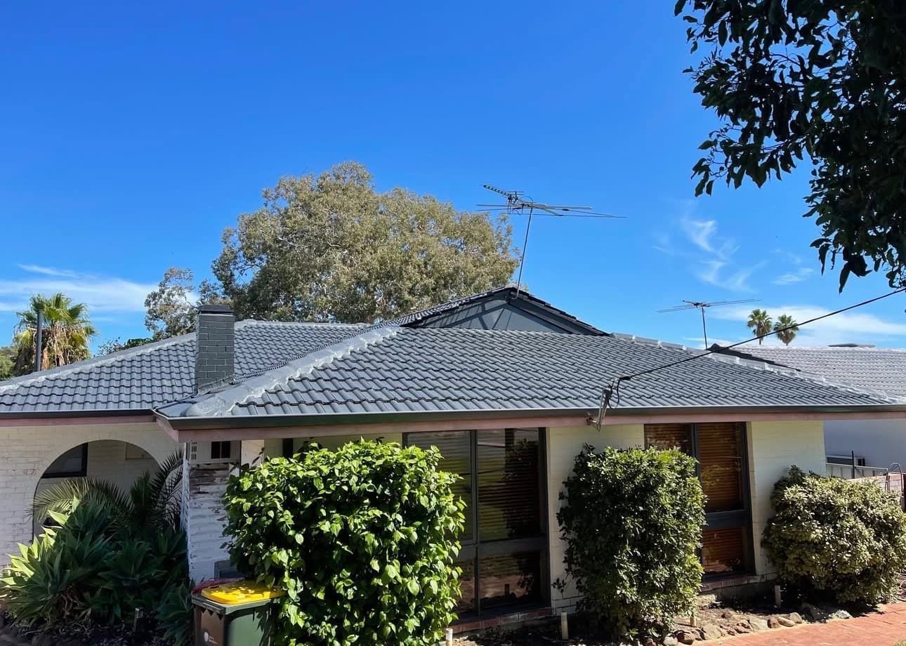 A single-story house with a grey tiled roof, off-white walls, and large windows, surrounded by green shrubs under a blue sky.