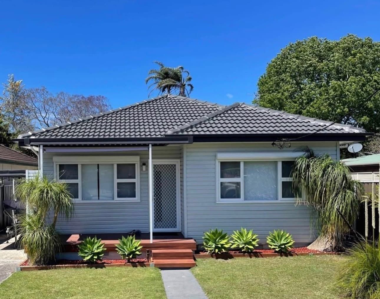 A single-story, light grey cottage with a dark tiled roof, a covered porch, and a small front lawn under a blue sky.
