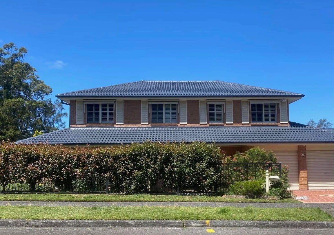 A two-story brick house with a blue tiled roof, fronted by a large hedge and a green lawn under a clear blue sky.