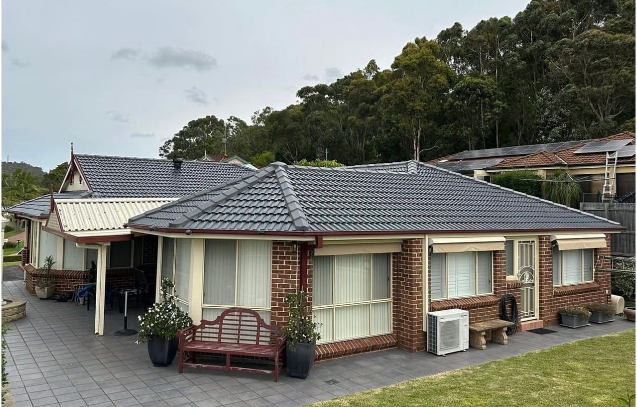 A multi-story brick house with a brown shingled roof, set against a background of green trees and a blue sky.
