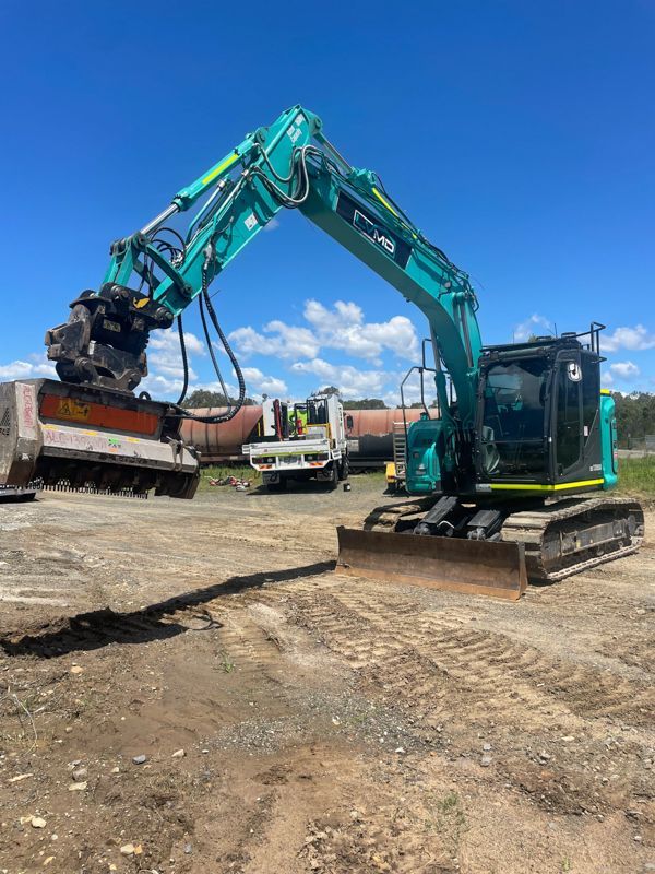 A teal excavator with a mulcher attachment, working on a dirt lot under a blue sky.