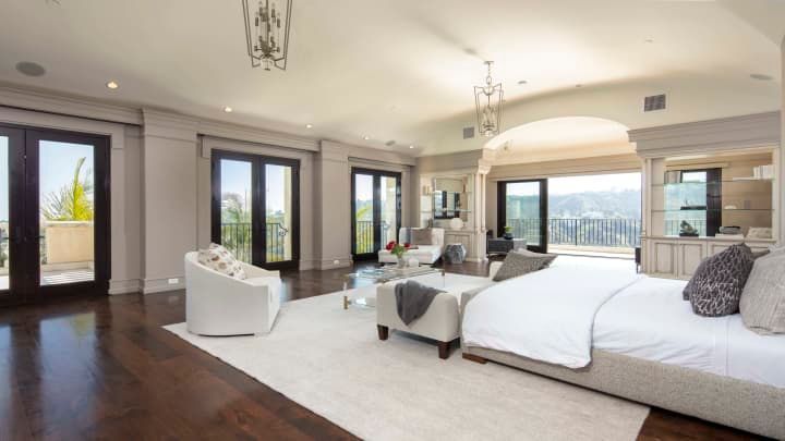 A spacious, modern master bedroom with dark wood floors, white furniture, and a large balcony overlooking the hills.