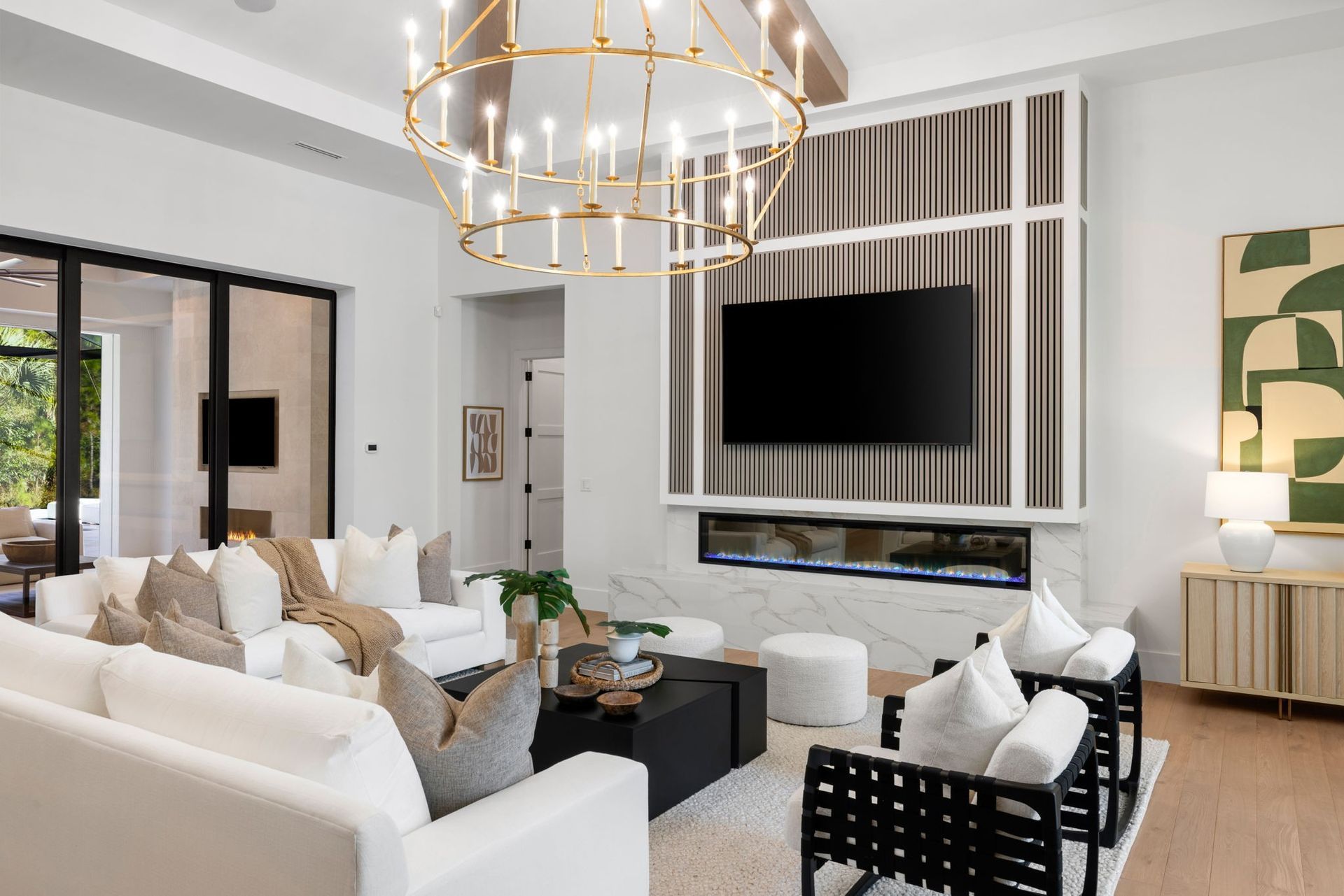 A bright, modern living room with white sofas, a large tiered gold chandelier, a fireplace, and a mounted television.