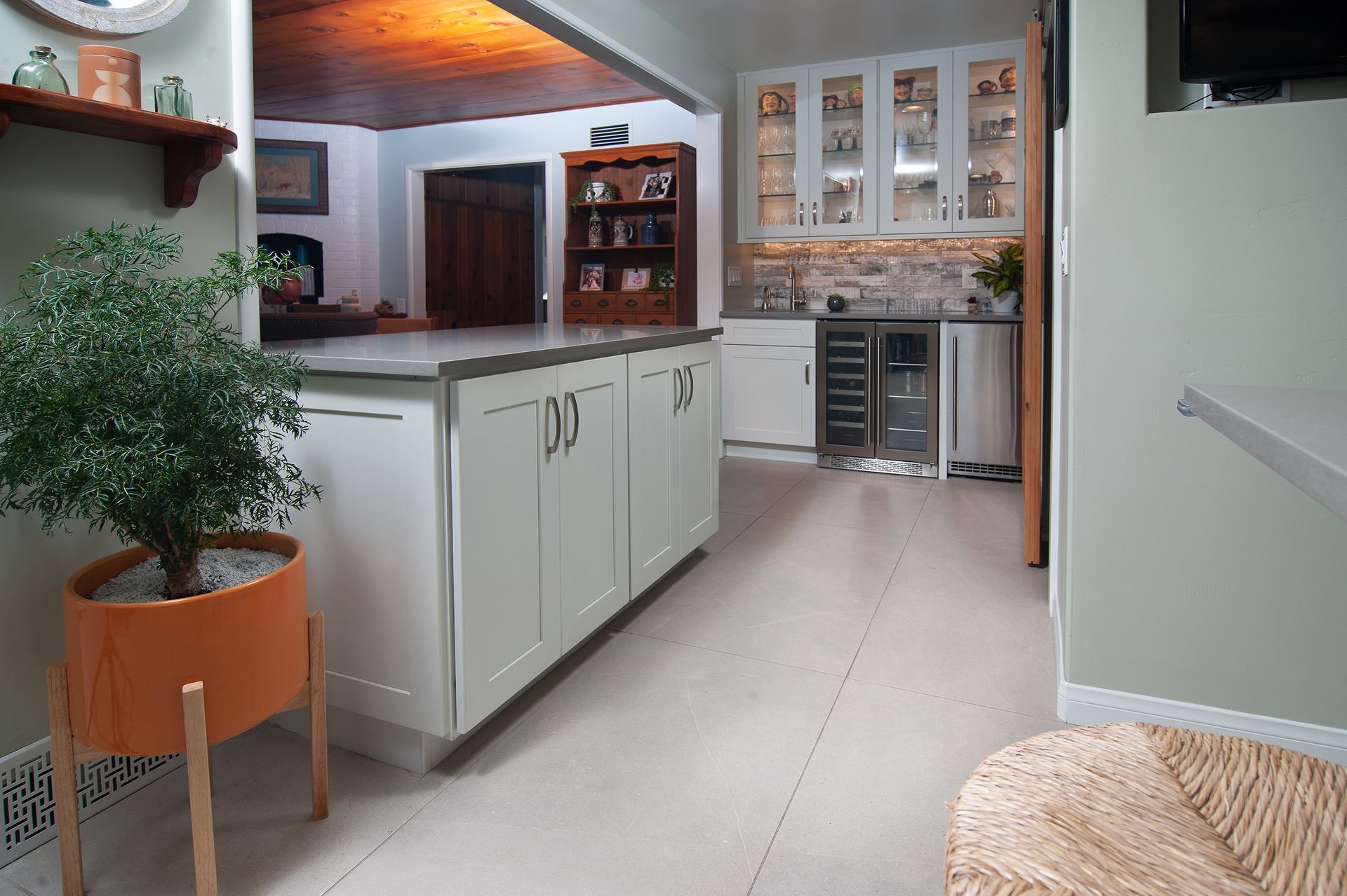 Kitchen with island, cabinets, built-in wine cooler, and a plant in an orange pot; view to a living area.