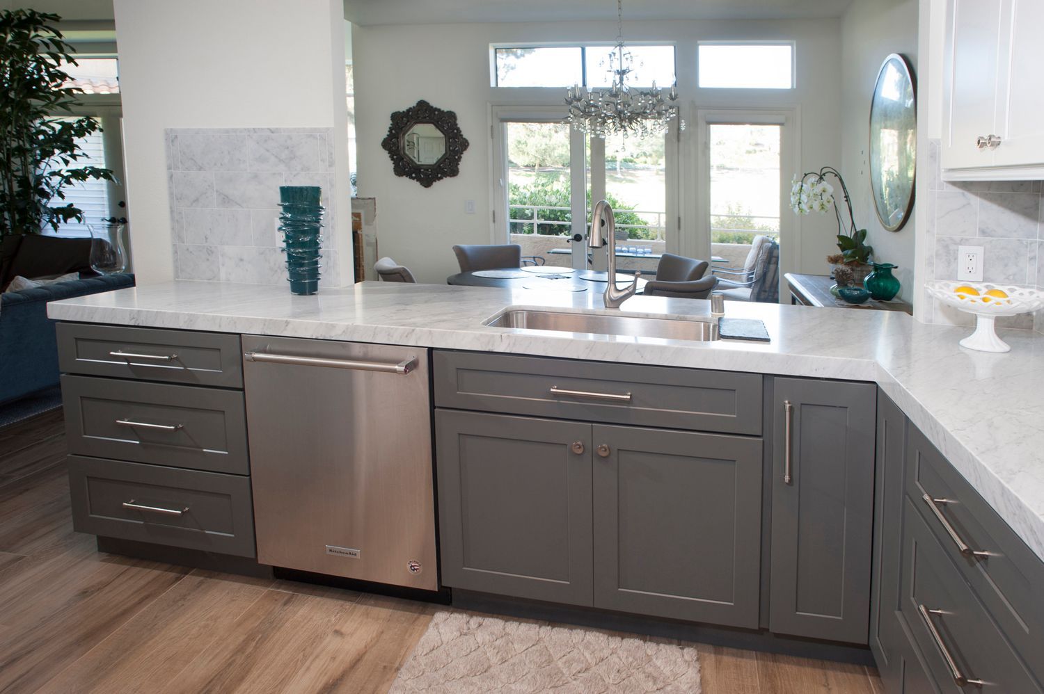 Kitchen with gray cabinets, stainless steel dishwasher, white countertops, and a view of a living area.